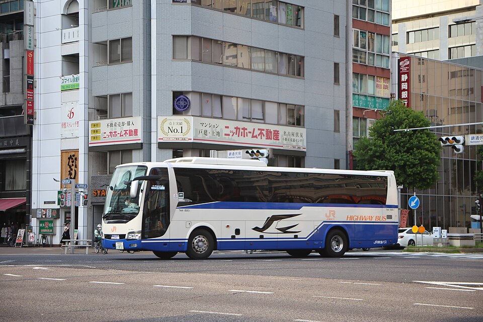 A white and blue JR highway bus, with HIGHWAY BUS written on its side, driving along a city street past office buildings